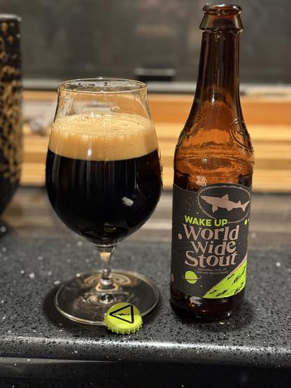 That snifter glass again, dark beer, tan head, and a brown bottle with a black labels there is a silver dogfish silhouette and a lime green triangle and the words “wake up”