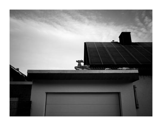 A black and white photo of a garage. On the edge of the roof is a plastic reindeer seems to run from right to left. In the background, a house roof with solar panels is visible.