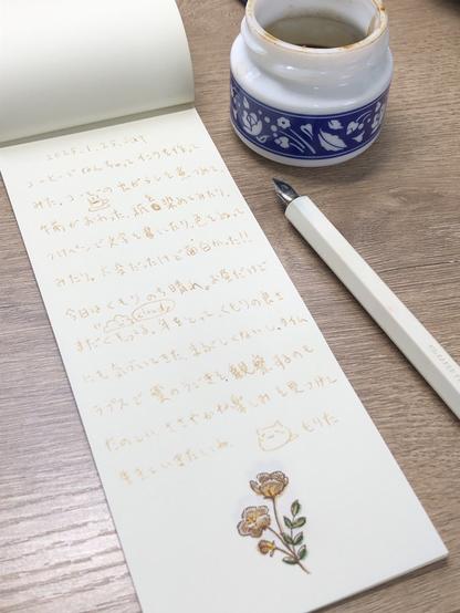 メモ帳、インクを入れた瓶、つけペンの写真。メモ帳にはコーヒーインクで書いた文章。