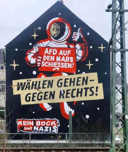 Streetartwall. A mural with an astronaut and an invitation to vote was sprayed/painted on the outside wall of a two-storey building.  The background is black with golden stars. Flying in the middle is a pictogram-like painted astronaut wearing a helmet and holding his thumb up. In front of him is a sign with the words: "Shoot the AfD on Mars" in German. Underneath is a large banner with the text: "Go vote, against the right!" And a little below: "Kein Bock auf Nazis". The mural refers to the 2025 federal elections in Germany and the warning against the right-wing Nazi party AfD, which is supported by Elon Musk. 
Info: The "Kein Bock auf Nazis" (KBaN) initiative (translated: "Does not like Nazis") is the largest independent youth initiative against right-wing extremism and racism in Germany. It raises awareness and networks with campaigns and concerts.