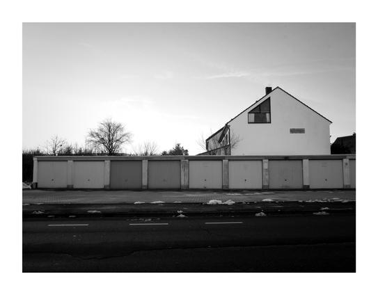 A black and white image of some garages with a large empty space in front of them. Some garage doors are dirty and they obviously have different color tones. There is no sign of life in the image.