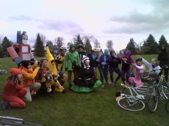 People in colourful costumes sitting on a grassy field around some bikes. Prominently on the left is one person dressed as Optimus Prime, the Transformer. Their costume is made of large cardboard boxes, too big for bike riding, so they came on a skateboard. There are also people dressed as a banana, Little Bo Peep, Bill Watterson's Calvin (with a stuffed Hobbes), and Barney the dragon, among others.