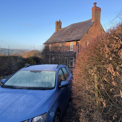 an old red brick cottage with two chimneys and grey slate roof, blue sky behind,  red-bronze hedge on one side, red-bronze trees on the other, with hills in the distance. In the foregound is a blue estate car with white icy windscreen. Red, white and blue.