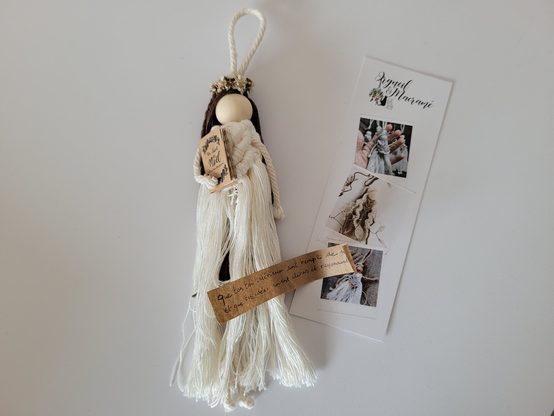 Petite poupée dont la robe blanche est réalisée avec la technique du macramé. L'objet a des tons blancs, ocres, et marrons. Elle a une longue chevelure brune et tient un petit livre.

Posé sur la poupée, un petit papier marron déroulé, où on lit : "Que ton toi intérieur soit rempli de sérénité et que tes idées soient claires et rayonnantes".

A côté, un marque-page indiquant "Orgueil & Macramé" avec trois petites photos d'autres réalisations de poupées de sortilège en macramé.