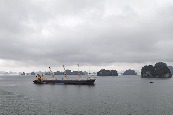 We're at sea, in a bay filled with limestone islands. The water is flat and grey, reflecting the heavy clouds that smother the sky. Some of the nearer, lumpy islands of rock show some detail of rough sides topped with dense vegetation but most of the dozens of islands we can see are little more than silhouettes becoming paler with distance, their shapes like rows of rotted teeth. A cargo ship sits at anchor in the middle of view. It has a long body, dark blue in colour, and a rectangular bridge block at its aft on the left in a light yellow. There is no sign of cargo on its deck but there are four tall cranes equally-spaced and ready to assist loading and unloading.
