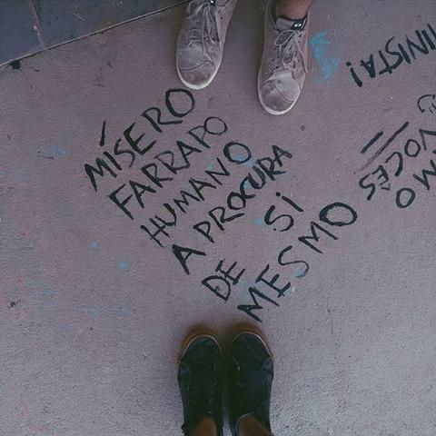 A person's feet are positioned above ground art featuring lyrics from O Terno's song 'Tudo Por Ti' (Everything For You). The visible words, written in Portuguese on a paved surface, are 'Mísero farrapo humano a procura de si mesmo' ('Miserable human rag in search of itself'). The image combines street art with a reference to Brazilian music, creating a unique visual and cultural statement.