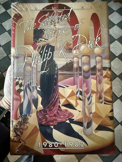 The cover of Volume 6 - The Selected Letters of Philip K. Dick - 1980-1982. It features a masked and head-dressed woman wearing a stars and moons trimmed robe that fades from black at the top to dusky pink at the bottom. She is standing in an abstract landscape of polygonal structures on a twisted checkered floor and strange pearl topped glass pillars that are kind of supporting an arched and domed ceiling. The woman seems to be performing a ritual or rite and has her hands raised. Five birds seem to be flying from her hands from which a strange light is emanating. There are mountains and a beach in the background. In the foreground bottom left, there are a couple of capuchins and a bird.