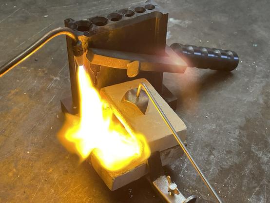 A burner ignites flames near the kouille resp. Casting mold for wire, which is fastened with a vice. The structure is located on a workbench, on which the tools and devices for metal melting are presented. There is 925 silver in the melting pot. 

Ein Brenner entzündet Flammen in der Nähe der Kokille bzw. Gussform für Draht, die mit einem Schraubstock befestigt ist. Der Aufbau befindet sich auf einer Werkbank, auf der die Werkzeuge und Geräte für das Metallschmelzen präsentiert werden. Im Schmelztiegel befindet sich 925 Silber.
