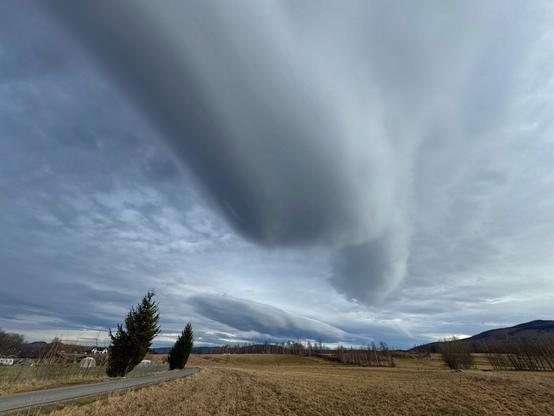 This image shows a landscape with an expansive sky featuring large, elongated lenticular clouds. These clouds have an unusual, smooth, lens-like shape. The scene includes a rural area with fields, a few trees, and distant hills. The lighting appears overcast, contributing to a dramatic and serene atmosphere.