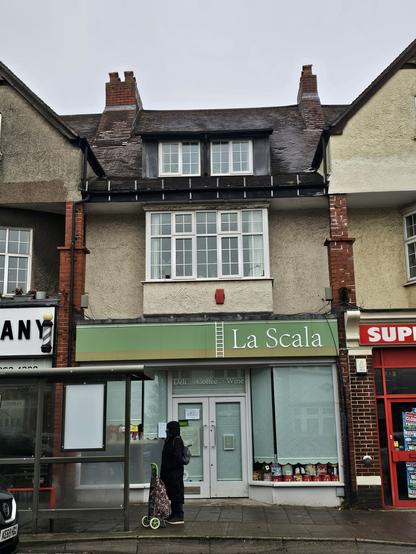 Photograph of a shop front, with grey blinds pulled down. The shop sign above the window reads 'La Scala' in mixed case serif. It is a delicatessen