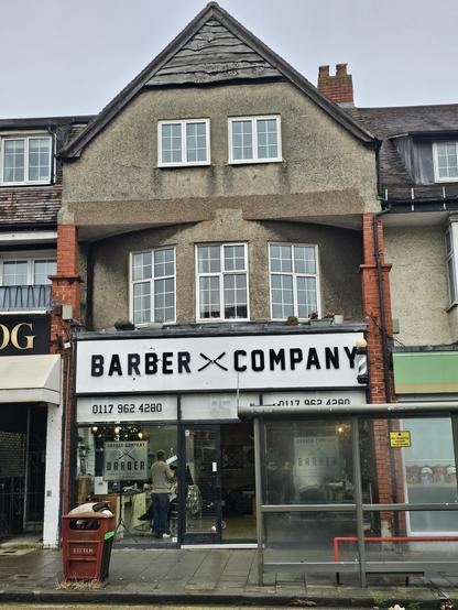 Photograph of a shop front, partially obscured by a bus stop. The shop sign above the window reads 'Barber Company' in ALLCAPS san serif.