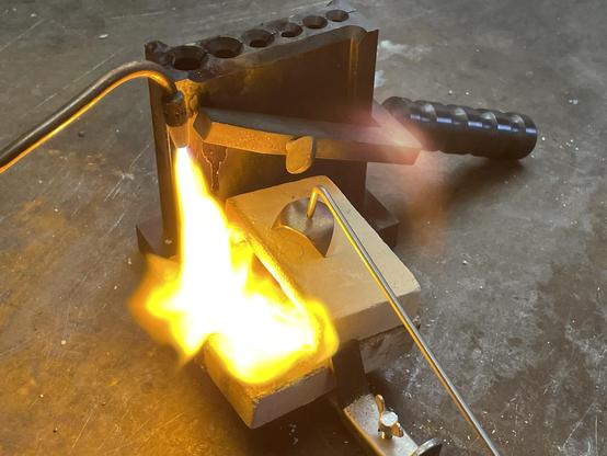A burner ignites flames near the kouille resp. Casting mold for wire, which is fastened with a vice. The structure is located on a workbench, on which the tools and devices for metal melting are presented. There is 925 silver in the melting pot. 

Ein Brenner entzündet Flammen in der Nähe der Kokille bzw. Gussform für Draht, die mit einem Schraubstock befestigt ist. Der Aufbau befindet sich auf einer Werkbank, auf der die Werkzeuge und Geräte für das Metallschmelzen präsentiert werden. Im Schmelztiegel befindet sich 925 Silber.