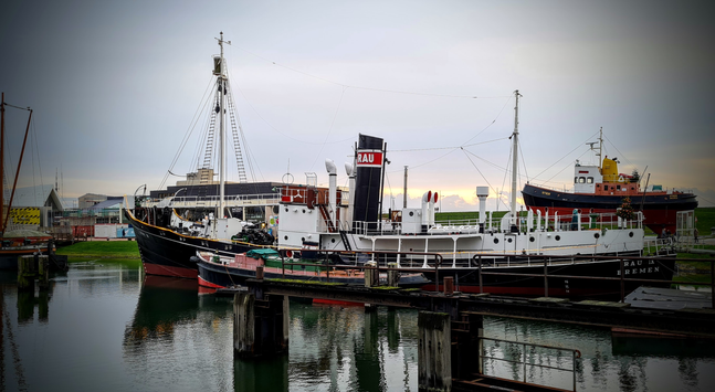 Ein alter Schiff liegt in einem Hafen. Weiter Schiffe sind festgemacht oder aufgedockt.