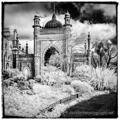 A toned black and white infrared photo of a gatehouse built in Mughal style with the roadway bordered by various exotic plants.