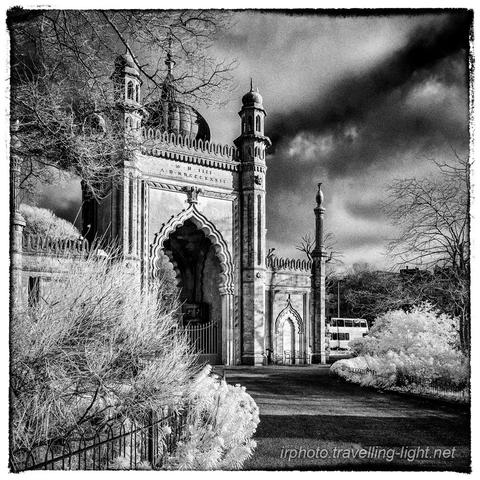 A toned black and white infrared photo of a gatehouse built in Mughal style with the roadway bordered by various exotic plants.