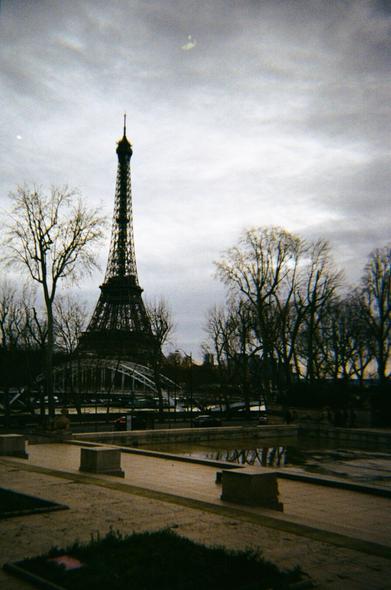 "View of the Eiffel Tower on a cloudy day, framed by leafless trees. A bridge is visible in the foreground, along with a reflective pool and paved pathways. The scene has a moody, overcast atmosphere." (AI generated text)