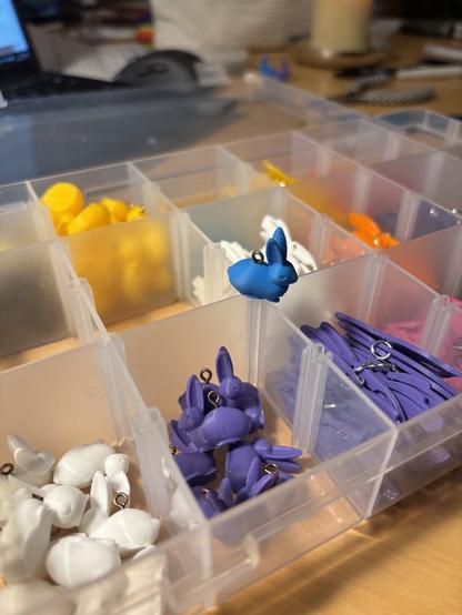Kunststoffkiste mit Fächern. Auf der Kante steht ein kleiner 3d gedruckter Hase in blau mit einer Öse am Rücken. In den Fächern sind weitere Hasen in lila und weiß zu sehen. Die Hinteren Fächer sind ebenfalls gefüllt. Es ist nicht zu erkennen mit was genau.
Im Hintergrund ist das Chaos eines Schreibtisches zu erahnen.