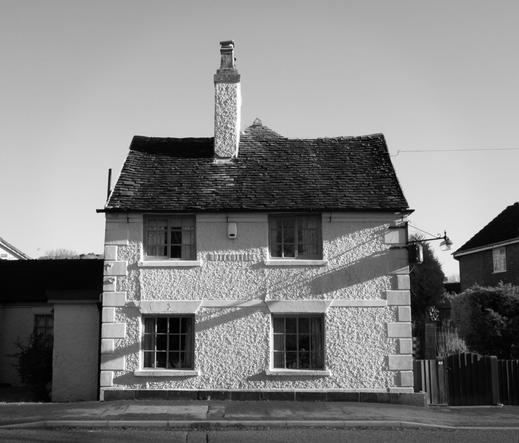 An old cottage, the tiled roof is a bit wonky, on the corner there is a street lamp attached to the house and the shadows from it and its electric box are cast across the whole front of the building.