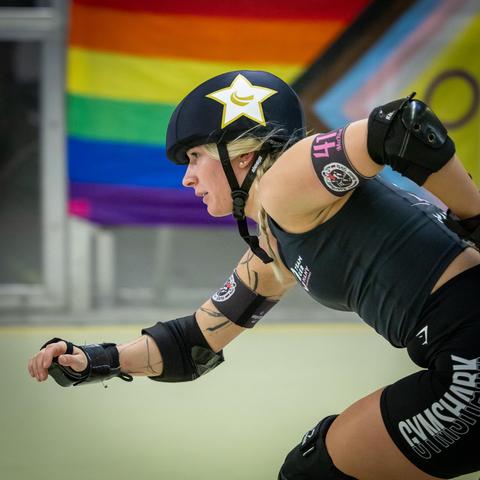 Color picture of a Roller Derby jamming person skating low in front of an LGBTQ+ Flag