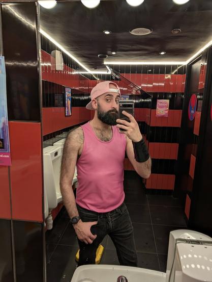 A selfie of a bearded man in a bathroom mirror. He's wearing a backward pink cap that says "brain not included", a pink leather tank top and black jeans. You can see body hair on his chest and shoulders. The bathroom has black and red tiling.