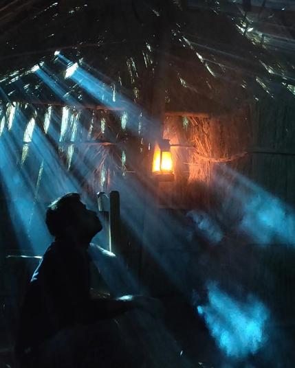 Cinematographer Abhishek Modak is completely silhouetted against beams of sky-blue light that penetrate the roof of a darkened grass hut, lit within only by a dim amber lantern. He is seated on the floor looking upward, as though deep in thought.