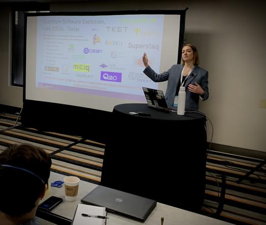 Kate Smith standing at lecturn giving a talk, with slides showing the logos of lots of quantum companies..