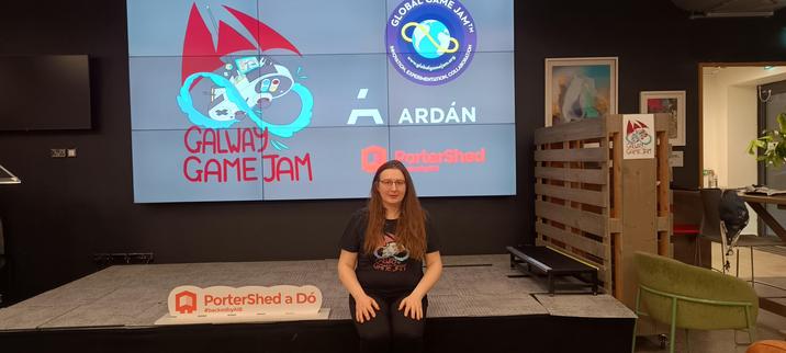 A white woman (me) sitting on the edge of a small stage with a video wall behind. The video wall is displaying the logos of Galway Game Jam, Global Game Jam, Ardán and Portershed. There is a movable barrier made of wooden palettes on casters on to the right of the stage, with a small green armchair in front, the armchair is facing towards the front of the building. One of the entrances/exits of the building is in the far right of the picture. There are some chairs and a desk at the entrance.

I am wearing black jeans and a black T-shirt with the Galway Game Jam Logo. I have long brownish blond hair and I am wearing glasses with a red frame.