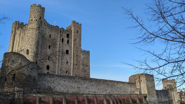 A view of Rochester Castle, with part of the outer wall in the foreground with the tall keep behind it. The keep isn't ruined, but it's very weathered and appears hollow. Three of the corners have square towers, while the fourth is round.