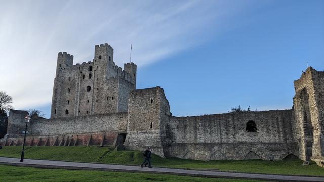 A view of Rochester Castle, with part of the outer wall in the foreground with the tall keep behind it. The keep isn't ruined, but it's very weathered and appears hollow. Three of the corners have square towers, while the fourth is round.