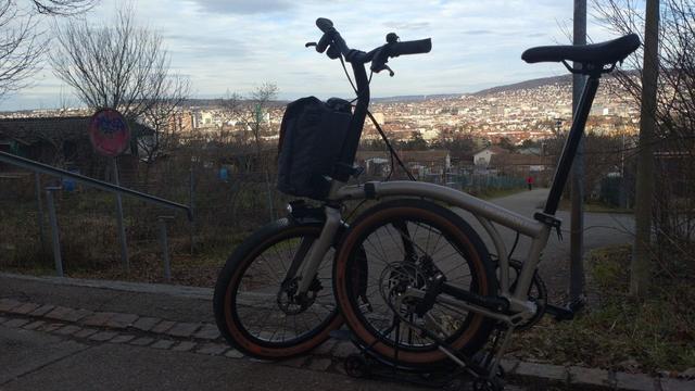Ein halb gefaltetes, graues Brompton Electric G-Line steht vor einem im schatten liegenden Treppenabgang. Im Hintergrund sieht man Zürich im Sonnenschein.