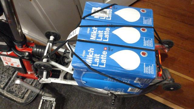 6× 1 Liter H-Milch mit den zwei Doppel-Gummistrapsen des Brompton-Gepäckträgers auf selbigen festgeschnallt. Das Rad steht in der Wohnung halb auf einer Schuhabtrofschale.