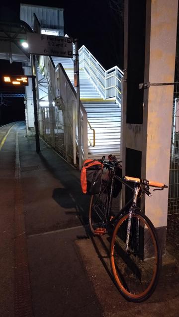 Orange single speed bike with panniers sitting on a train station platform at night