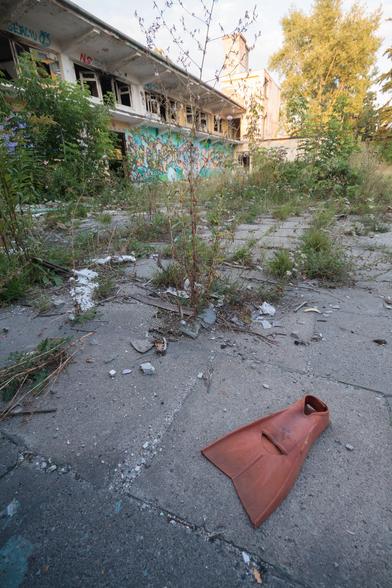 A red-brown swimming flipper lies on rough concrete punctuated by foliage - in the background, a derelict two-storey building glows in evening sunlight.