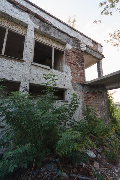 Outside an abandoned, two-storey building, walls covered in an ornate random pattern of small, curved fragments of white ceramic - which has fallen off in large areas to reveal rough red brickwork and concrete blocks. Glassless windows reveal darkened rooms inside; in the foreground at ground level is dense green foliage growing out of loose rubble.