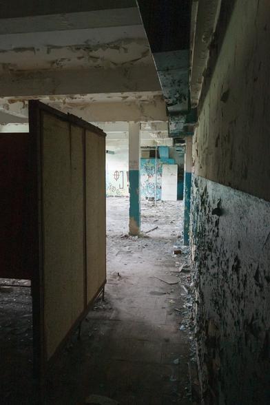 Inside an abandoned building - an empty room with peeling paint, sparse graffiti and a rough tiled floor, with wooden cubicle walls for changing in. Diffuse evening light spills in from the background - the foreground is in partial darkness.