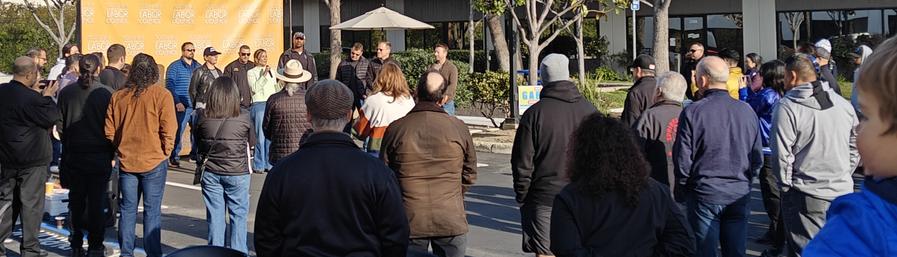 An outdoor rally with a woman speaking at a handheld microphone surrounded by a listening crowd.