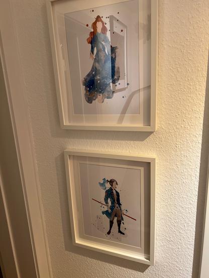 Two prints mounted on a wall in frames. One shows a red-haired woman holding out her hand, the other a young man with a spear and a glowing spirit on his shoulder.