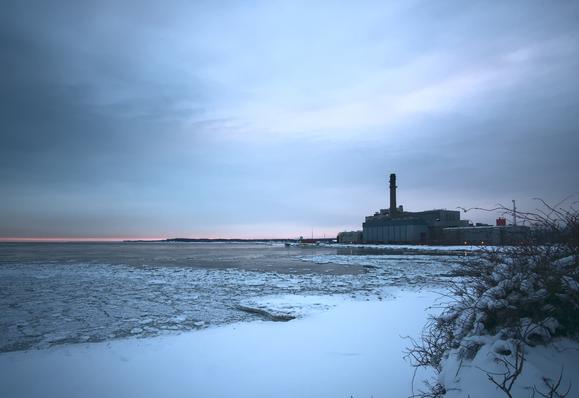 Photo taken from a slight rise overlooking Dunkirk bay in rural western NY state. Snow in the foreground gives way to broken ice on the surface of a dark lake above, fading into dark water beyond, and a slice of land in the distance extending from the right. Also on the right: a one-smokestack edifice which I believe is a decommissioned fossil-fuel power plant. Its main smokestack rises at least 100 feet above the rest of the massive pile. The sky above all this is striated, blended gray/blue/white, with a strip of peach on the horizon, like a gen Z hair dye job faded halfway.