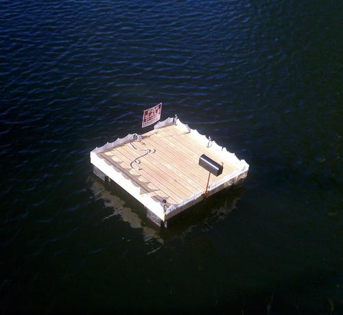 A square dock about 6 feet to each side is floating in the water and framed by a white picket fence. It has a mailbox on a post on one side, and a gap in the fence on the other side where a For Sale By Owner sign is posted.
