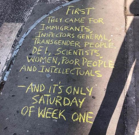 A photo of a sidewalk with a political message written in yellow chalk. The text reads, "First they came for immigrants, inspectors general, transgender people, DEI, scientists, women, poor people, and intellectuals—and it's only Saturday of week one." Sunlight and shadows partially fall over the chalk writing, emphasizing its impromptu and grassroots nature. The message echoes concerns about early actions during a political administration's term, highlighting marginalized groups. The setting is urban, with a slightly worn concrete pavement surface.