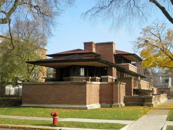 The image showcases a distinctive architectural structure characterized by its horizontal lines, flat roof, and extensive use of stone and brick materials. The design reflects elements of the Prairie School architectural style, typically associated with Frank Lloyd Wright. It features large overhanging eaves and a strong connection to the surrounding landscape, with an emphasis on integrating the building into its environment. The setting appears to be a residential area, with trees and a sidewalk visible in the foreground.

Image Credits: Wikimedia / Teemu08 / CC BY-SA 3.0