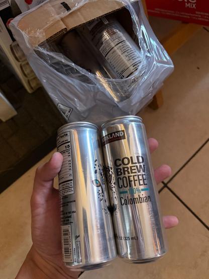 A pair of Kirkland Signature brand cold brew coffee cans, held in my hand. A carton of more coffee cans can be seen behind.