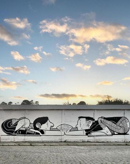 Streetartwall. A great black and white mural with two reclining figures was sprayed/painted in a simple but detailed comic style on a long gray street wall. The background above the wall is a blue, cloudy sky. The mural shows a girl and a boy in different black and white clothes, lying on the ground facing each other with their hands tightly intertwined. Their posture is unusual. Both are sitting on their buttocks, their legs bent flat and their arms stretched out towards each other on the floor. A kind of yoga session for lovers.
Info: The mural was created during the G20 meeting and follows the poetic approach that it is always possible for different characters and lifestyles to come together.