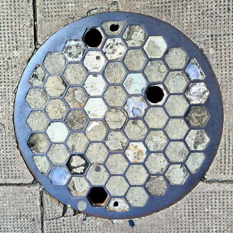 A metal access cover filled with hexagonal cells. Most of these have been filled with concrete, but a few of the original glass prisms remain in place.