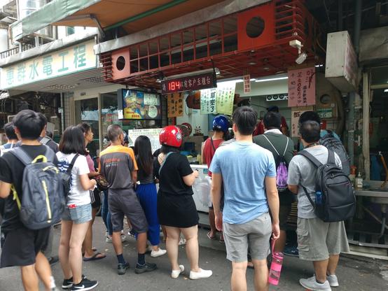 A tea shop on a street in Taipei, with a queue of people who are waiting to order or have already ordered their bubble tea. Above the counter there is a number display in LED matrix style that shows the number 184, and various signs and hand-written posters that show Chinese characters are attached to the facade above the crowd. It is warm, many are wearing short pants, and there is a relaxed atmosphere to it.