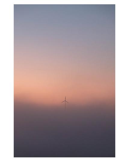 Foto im Hochformat. Sonnenaufgang, alles ist neblig. Darüber ist der Himmel, am Horizont rötlich, darüber hellblau. Das einige, was im Nebel zu sehen ist, ist der obere Teil einer Windkraftanlage. Einer der drei Rotoren des Propellers zeigt genau nach oben.
