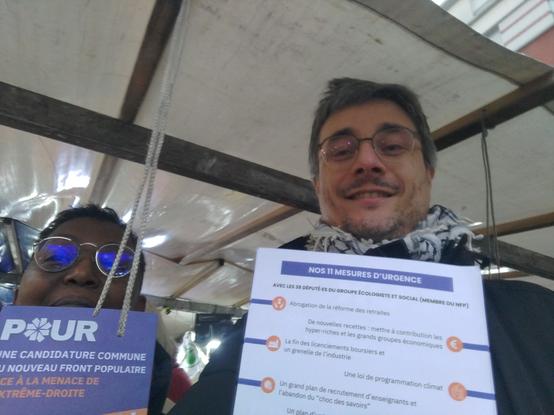 Deux personnes sur un marché montrent un tract. on lit en titre du recto "Pour une candidature unique du Nouveau front populaire face à la menace de l'extrême-droite. Au verso, " Nos 11 mesures d'urgence ".