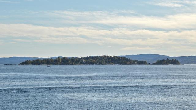 A photo of an small island in the Oslofjord. There are hills in the distance and the sky is a hazy blue with white clouds in it.