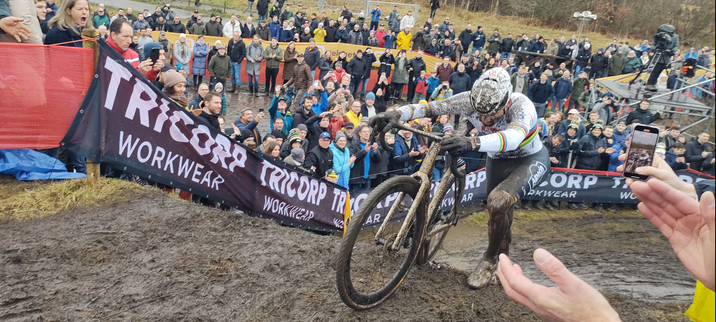Cyclocrosser in rainbow jersey pushing his bike up a ramp. Spectators are cheering behind banners.