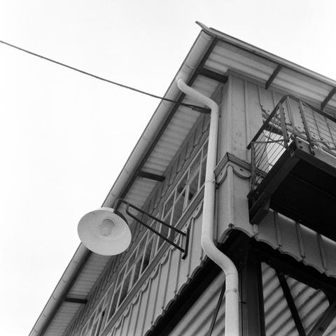 A black and white photograph taken from a low vantage point, focusing on the corner of a wooden building. The building's roof, which casts a shadow over the lower portion of the image, is equipped with a gutter and downspout. A small, round, white light fixture is attached to the building's exterior wall beneath the roofline. A metal balcony, complete with intricate metal railings, extends from the second floor. The building's facade is composed of vertical wooden slats, creating a striped appearance. A single electrical wire diagonally crosses the top of the image, partially obstructed by the roof. The background is a solid, light gray, indicating a clear, overcast sky.